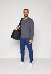 Grauer Kapuzenpullover mit "Champion"-Logo, blaue Jogginghosen, weiße Sneakers und eine schwarze Sporttasche. Einfaches Design, lässige Passform und einfarbige Farben.