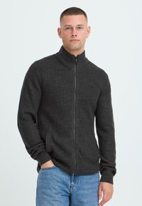 Mørkegrå strikket zip-up genser med strukturert stoff, stående krage og sidelommer, sammen med lyseblå jeans.
