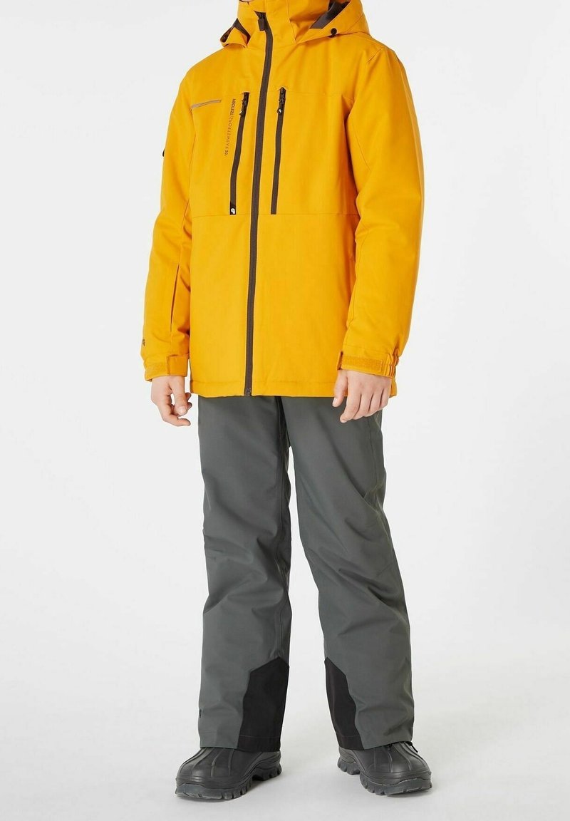 Veste imperméable jaune vif avec poches zippées, associée à un pantalon gris avec des accents noirs et des bottes noires robustes, conçue pour une utilisation en extérieur.