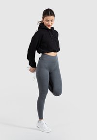 Mustassa cropped-huppari, jossa on nyöri, yhdistettynä harmaisiin ribattuihin korkeavyötäröisiin legginsseihin. Mallilla on valkoiset urheilukengät.