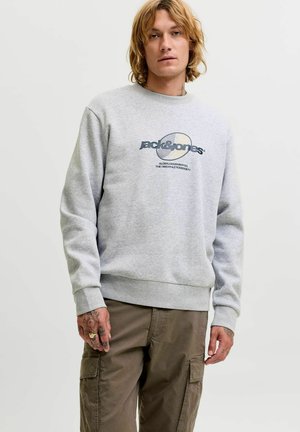 Personne aux cheveux blonds ondulés porte un sweat-shirt gris clair Jack & Jones et un pantalon cargo kaki, debout devant un fond uni clair.