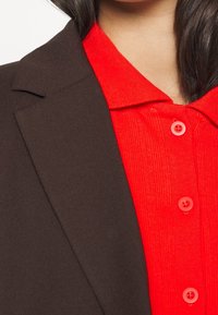 Brązowa marynarka z tkaniny z wyraźnymi klapami, zestawiona z teksturowaną czerwoną koszulą z ryftami i okrągłymi guzikami. Zbliżenie na obszar kołnierza.