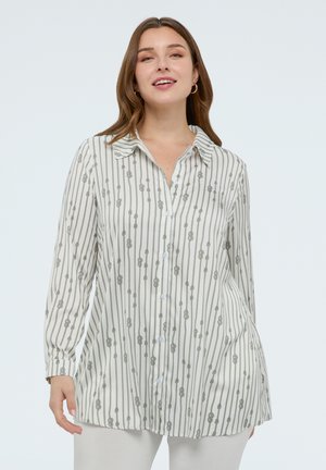 Mujer con una camisa blanca de manga larga abotonada, con rayas verticales y diseños de nudos, posando contra un fondo claro y liso.