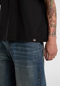 Side view of person wearing faded blue jeans and a black t-shirt with a small Dickies logo tag near the hem, showing tattoos on their arm.