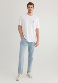 White cotton t-shirt with a round neckline, light blue graphic logo, paired with light blue denim jeans and beige sneakers.