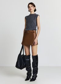 Haut sans manches en maille torsadée grise, mini-jupe en cuir marron, bottes brillantes noires montant aux genoux, et sac fourre-tout noir avec un accent doré.