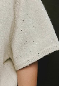 Cream knit sleeve with small silver sequin embellishments, showing part of a bare arm against a dark background.