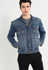 Jeans jakna u srednje plavoj boji s prednjim dugmadi, dva džepa na prsima i vidljivim šavovima. Klasičan dizajn s lagano izblijedjelom teksturom.