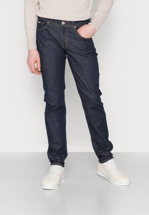 Dunkelblaue Jeans aus Denim mit geradem Bein, fünf Taschen und Kontrastnähten, kombiniert mit weißen Sneakers und einem leichten Pullover.