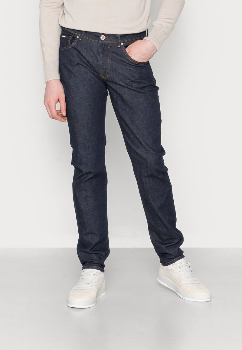 Jeans de mezclilla azul oscuro con corte recto, cinco bolsillos y costuras de contraste, combinados con zapatillas blancas y un suéter ligero.
