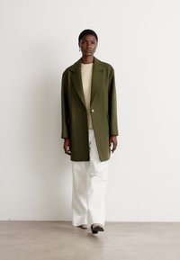 Oversized olive green blazer with notched lapels, button closure, layered over a beige top and white wide-leg trousers. Smooth fabric texture.