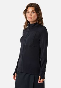 Vrouw met schouderlang bruin haar die een zwarte jas met rits en duimgaten draagt, staand tegen een effen lichte achtergrond.