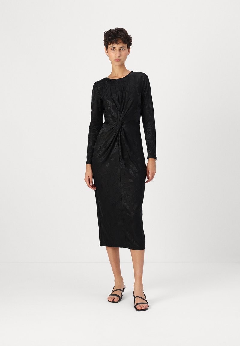 Vestido midi preto com mangas longas, detalhe frontal torcido, textura floral e decote arredondado. Usado com sandálias pretas planas.