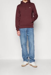 Sweat à capuche marron avec poche kangourou et capuche à cordon, associé à un jean bleu clair et des baskets blanches avec des accents beiges.