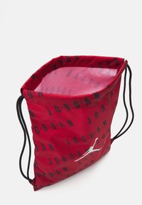 Crvena torba na izvlakač od sjajnog materijala s crnim natpisom "JORDAN". Ima bijeli logo akcent. Unutrašnjost je glatka i obložena.
