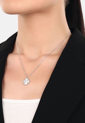 Silberne Halskette mit einem floralen Anhänger, der eine glatte, polierte Oberfläche und eine zarte Kette aufweist, getragen über einem schwarzen Blazer.