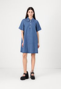 Denim kleita ar īsām piedurknēm, apkakli un priekšējo pogu aizdari, ar baltām vertikālām svītrām un neapstrādātu apakšmalu. Saskaņota ar masīvām melnām kurpēm.