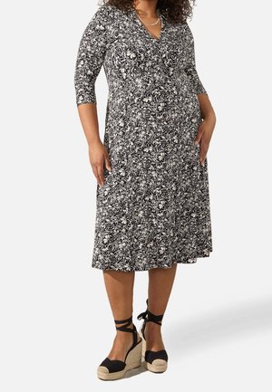 Vrouw draagt een zwart-witte bloemenjurk tot op de knie met driekwartmouwen en zwarte espadrille sleehakken met enkelbandjes.