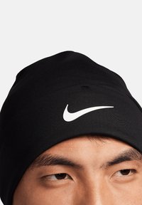 Berretto nero a maglia con una texture liscia, dotato di un logo Nike bianco sulla parte anteriore. Il design è aderente e presenta una parte superiore arrotondata.