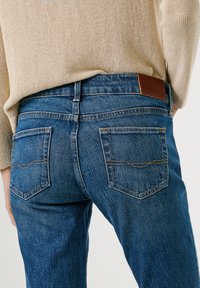 Modré džínsy z denimu s stredným pásom, piatimi vreckami a hnedou koženou nášivkou na chrbte. Prešití svetlohnedou farbou okolo vreciek.