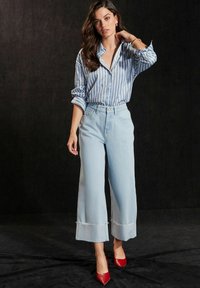 Jeans de pierna ancha en azul claro con dobladillo deshilachado, combinados con una camisa de botones a rayas en azul y blanco y unos tacones rojos de punta.