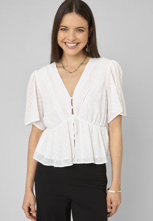 Femme souriante aux longs cheveux bruns portant un chemisier blanc à manches courtes boutonné avec taille péplum et pantalon noir sur fond gris.