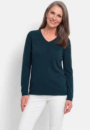 Femme souriante aux longs cheveux gris portant un pull vert foncé à col en V et un pantalon blanc, debout devant un fond uni.