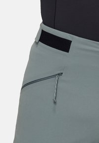 Pantalons gris clair avec une texture lisse. Présente une taille élastique noire, une poche zippée sur le côté et des détails de couture contrastants.