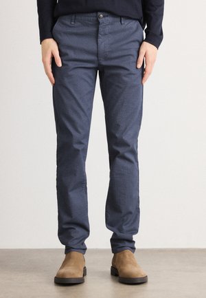 Navy blue slim-fit trousers made of textured fabric, featuring front pockets, a zip fly, and a single button closure. Worn with tan ankle boots.