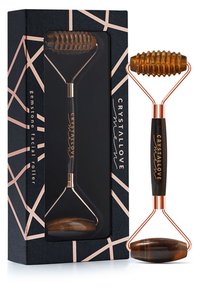 Roller do twarzy z kamieniami szlachetnymi z ciemnym bursztynowym kamieniem, metalowymi akcentami w kolorze miedzi i czarnym uchwytem, zapakowany w pudełko o geometrycznym wzorze.