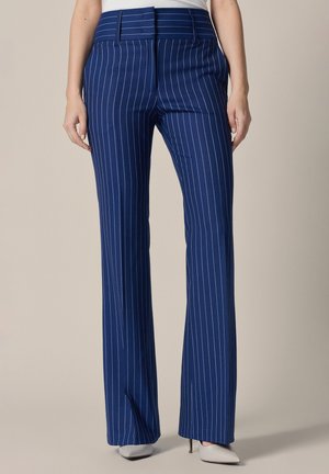 Granatowe, wąskie spodnie w prążki o wysokim stanie, noszone z białymi szpilkami z ostro zakończonymi noskami i włożonym do środka topem.