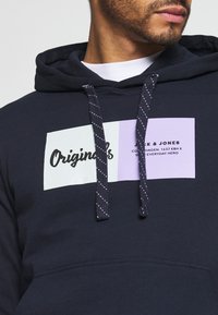 Marinblå huvtröja med dubbel patch-design med texten "Originals" i svart och en lila sektion med text. Inkluderar randiga dragsnören och framficka.