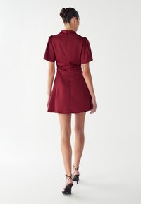 Aubergine satino suknelė su trumpomis pūstomis rankovėmis, aptempta juosmens dalimi ir plačia sijonu. Yra nugarinis užtrauktukas ir lygus paviršius. Juodos diržinės aukštakulnės bateliai.