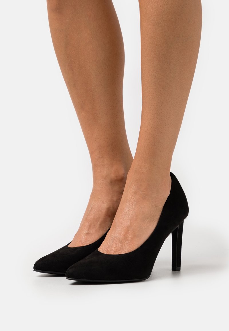 Beine, die klassische schwarze Wildleder-Pumps mit spitzer Zehenform und Stiletto-Absätzen auf einem weißen Hintergrund tragen.
