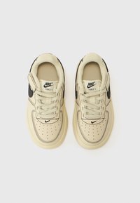 Bež Nike Air Force 1 tenisice imaju crne akcentne boje, perforirani nos i podstavljeni ovratnik. Dizajn uključuje vezice i potpisni logo.