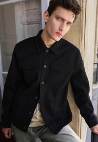 Jachetă neagră, cu nasturi, cu două buzunare pe piept, având o textură fină și un design structurat. Purtată peste un tricou cu guler rotund și dungi.