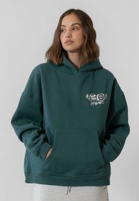 Sötétzöld kapucnis felső, pamut keverék anyagból készült, elülső zsebbel és két mesefigura grafikai mintával a mellkasán.