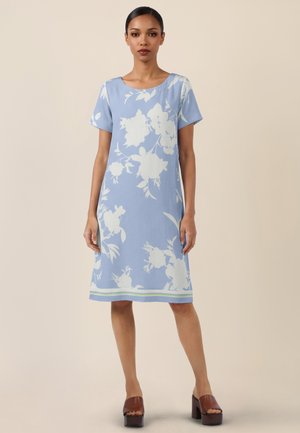 Frau trägt ein hellblaues knielanges Kleid mit weißen Blumenmustern und kurzen Ärmeln, kombiniert mit braunen Plateau-Absätzen.