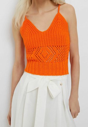 Frau trägt ein leuchtend orangefarbenes, gestricktes Crop-Top mit Spaghettiträgern und einen weißen, hoch taillierten Rock, der mit einer Schleife in der Taille gebunden ist.