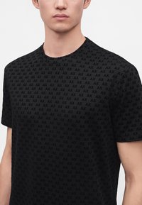 Jeune personne portant un t-shirt noir à manches courtes orné de petits logos "AX" répétitifs sur un fond uni clair.