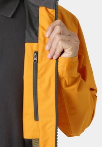 Ruka rozepíná jasně oranžovou bundu a odhaluje vnitřní kapsu se zipem na tmavé texturované košili.