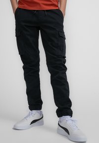 Zwarte cargobroek met een taps toelopende pasvorm, elastische cuffs, zijzakken en een verstelbare tailleband, gecombineerd met witte sneakers en zwarte accenten.