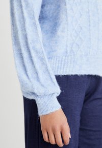 Hellblauer Strickpullover mit strukturiertem Muster, Ballonärmeln und geripptem Saum. Der Stoff wirkt weich und fluffig.