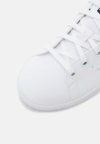 Sneaker bianco con tomaia in sintetico liscio, punta arrotondata e lacci piatti. Occhielli e sottili dettagli di cucitura sul lato. Suola bianca morbida.
