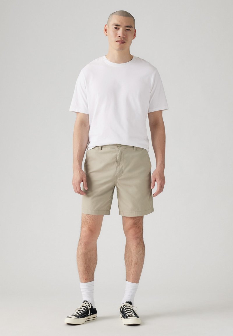 Pantaloni scurți bej cu față plată și bucle pentru curea, asortați cu un tricou alb simplu, cu mânecă scurtă. Tenisi negri cu talpă joasă și șosete albe.