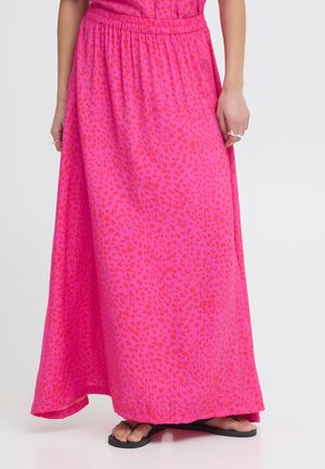 Frau trägt einen leuchtend rosa Maxirock mit einem dezenten roten Blumenmuster und schwarzen Flip-Flops, steht vor einem schlichten Hintergrund.