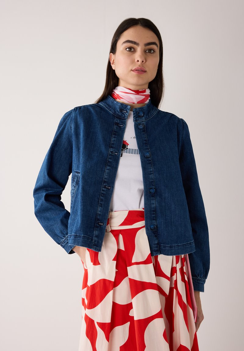Woman wearing a dark blue denim buttoned jacket, white shirt, red-and-white patterned skirt, and matching neck scarf against a plain background.