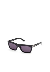 Gafas de sol rectangulares negras con lentes tintados en púrpura. Presentan un elegante marco de plástico y acentos metálicos en las patillas. Marcadas con "GUESS."