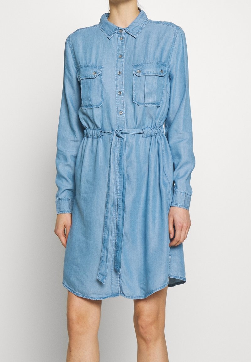 Robe chemise en jean bleu clair avec manches longues, deux poches poitrine, fermeture à boutons et taille ceinturée. Ourlet légèrement arrondi.