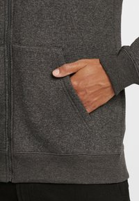 Siva zip-up dukserica s rebrastim manžetama i pojasom. Ima dva prednja džepa; ruka odmara u jednom džepu, pokazujući glatku teksturu tkanine.